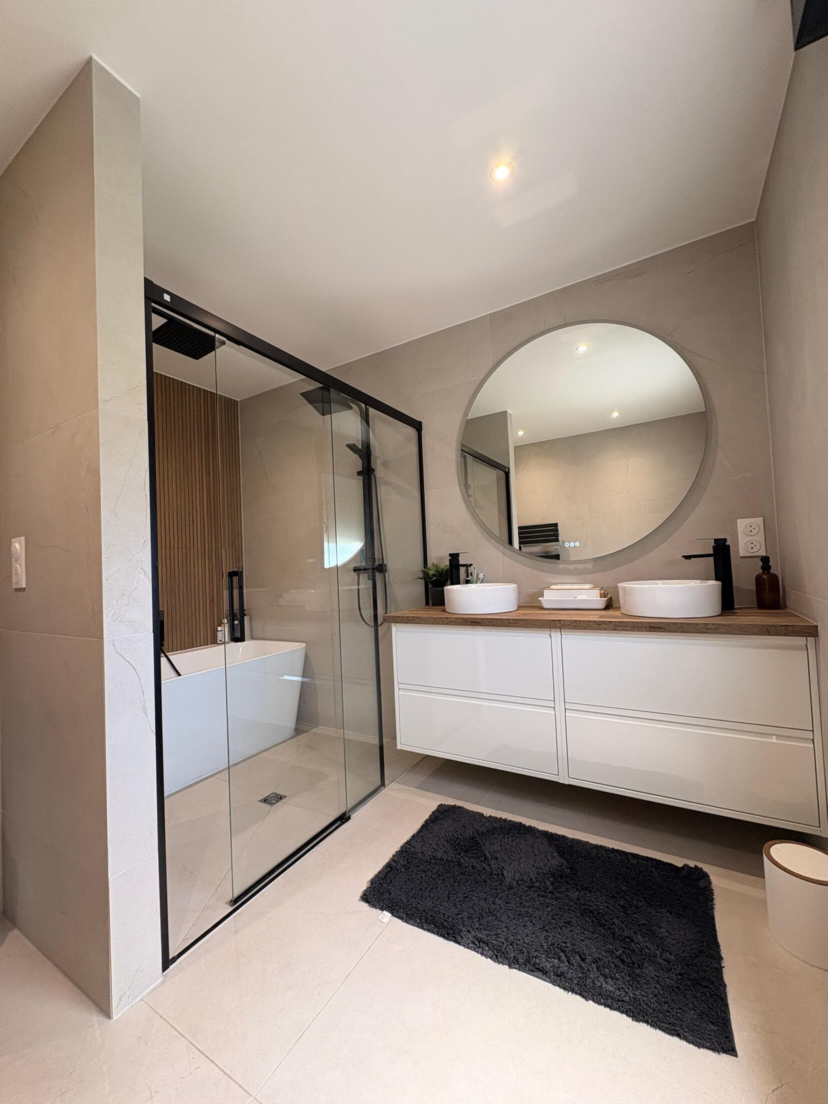 La salle de bain finie : douche vitrée, double vasque et miroir rond rétroéclairé